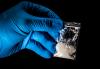 A hand with a blue rubber glove holding a transparent plastic sachet with a white powder